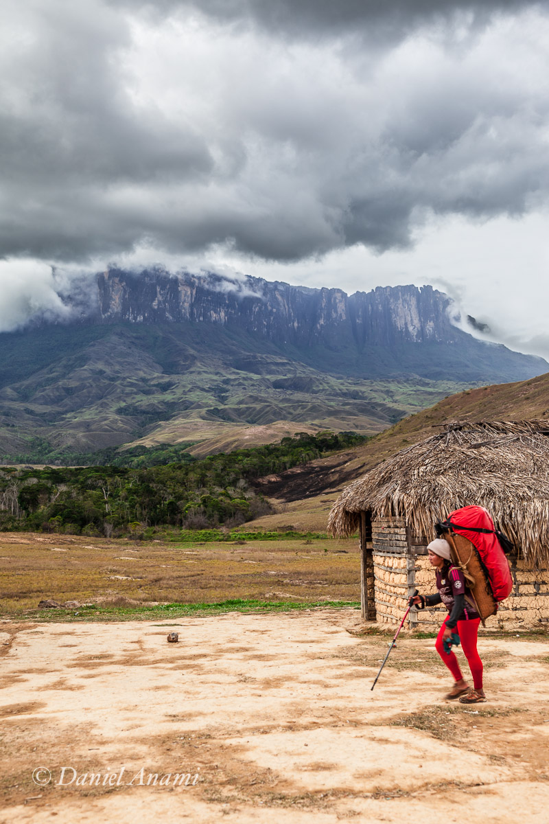 Porteadora com calça vermelha e grande carga, construção de pau a pique e palha e tepui Kukenán ao fundo. Nuvens carregadas no céu. Foto Daniel Anami, 13/03/2016.